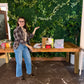 A woman wearing a plaid shirt and blue jeans stands next to a wooden table displaying various gift bags and packages, set against a green artificial ivy wall.