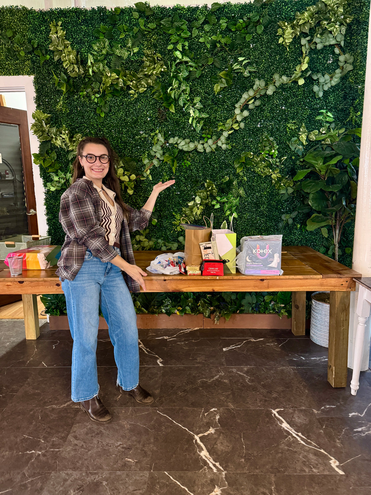 A woman wearing a plaid shirt and blue jeans stands next to a wooden table displaying various gift bags and packages, set against a green artificial ivy wall.