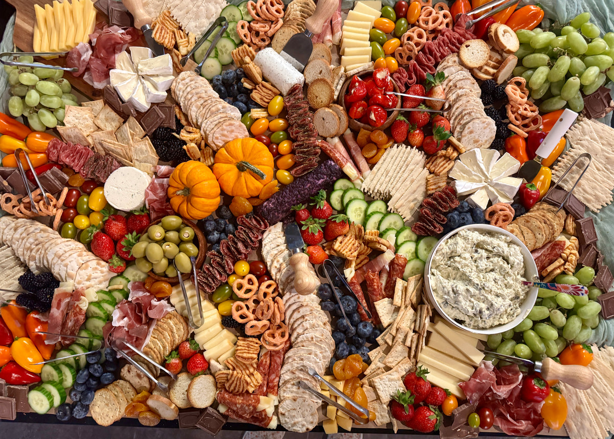 A large, beautifully arranged charcuterie board filled with sliced cheeses, crackers, cured meats, fruits, vegetables, pretzels, nuts, and a bowl of dip, photographed from above on a dark surface.