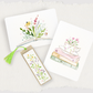 Three watercolor-style bookmarks or card-like stationery pieces arranged on a textured white background: an open book with wildflowers, a stack of books with a teacup and flowers, and a vertical floral bookmark with a green tassel.